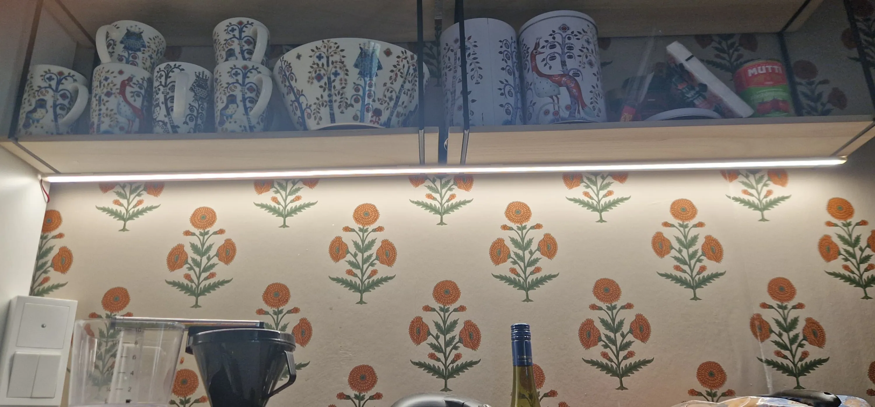 Keittiönäkymä, jossa vaalean puisen hyllyn päällä on valkoisia mukeja, kulho ja säilytysastioita sinisillä ja punaisilla puu- ja eläinkuvioilla. Oikealla hyllyllä on punainen Mutti-tölkki. Hyllyn alla oleva LED-valo valaisee seinää, jossa on toistuva oranssien kukkien ja vihreiden lehtien kuvio vaalealla pohjalla. Alhaalla näkyy valkoinen pistorasia, läpinäkyvä mittakuppi ja pullo.