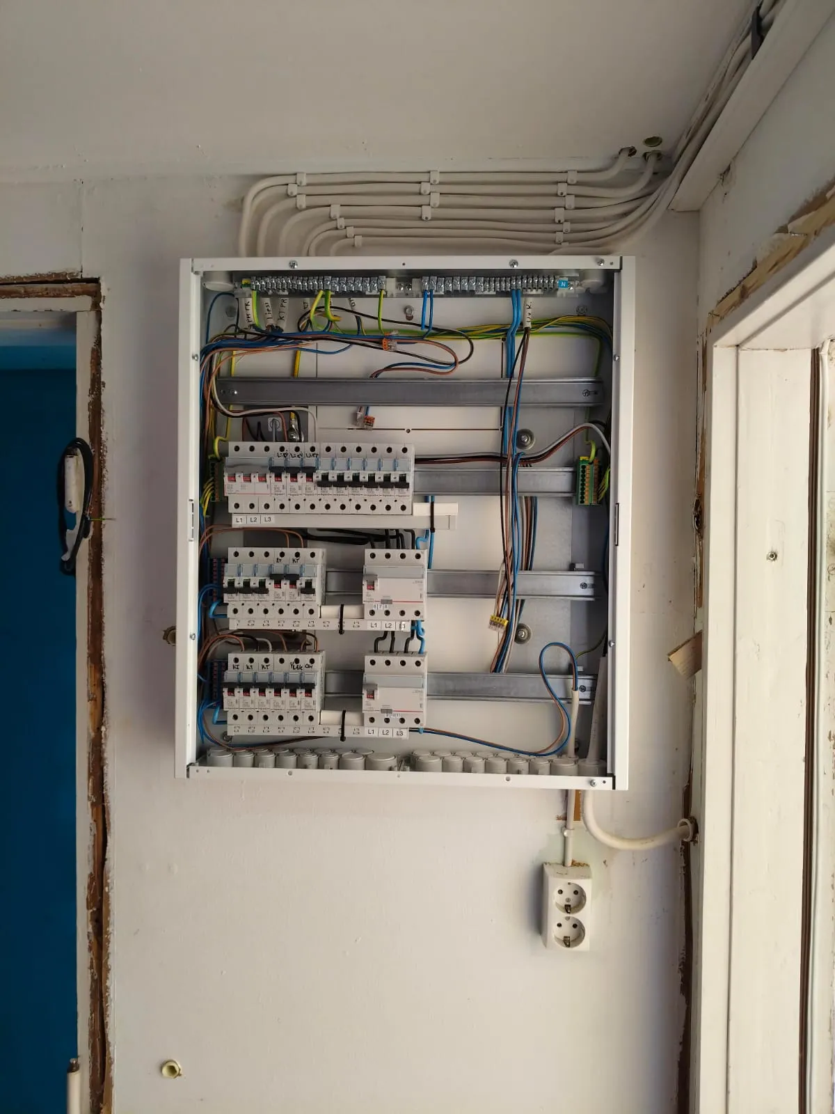 Avoin sähkökeskuksen kaappi, jossa näkyvissä johdot ja sulakkeet valkoisella seinällä.