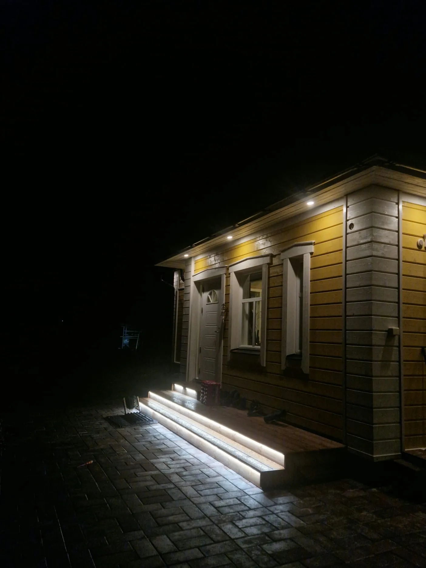 Keltaisen puutalon sisäänkäynti, jonka portaat on valaistu yöaikaan.